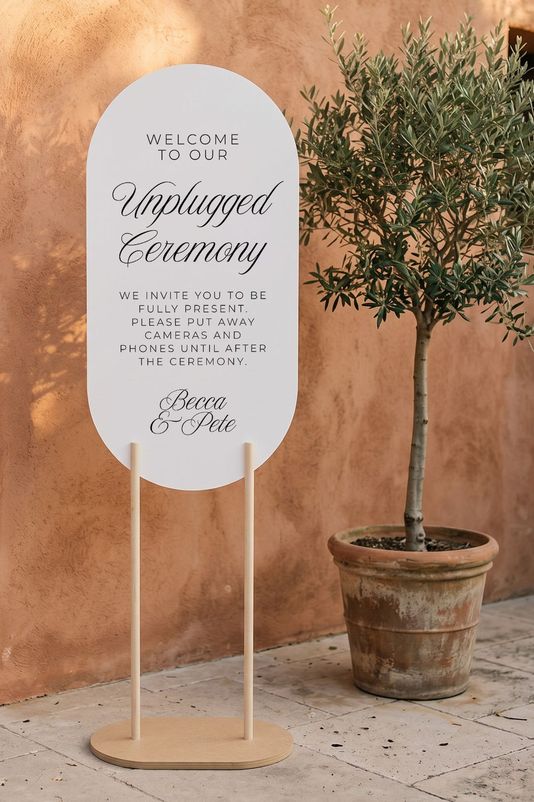 Oval unplugged ceremony sign beside an olive tree and terracotta wall #shape_Oval#size_Signature